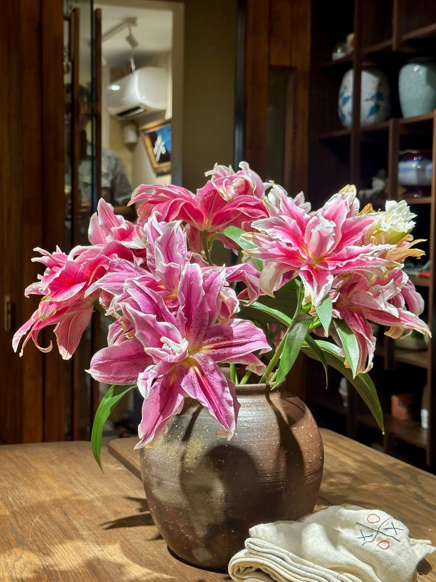 Pink double lilies in a rustic clay vase on a sunlit wooden table